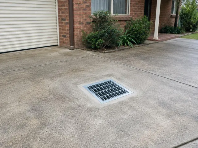Driveway drain grate set in concrete paving beside a brick house in Castle Hill